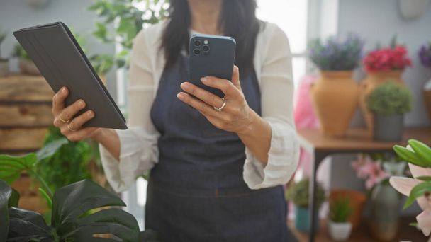 Una mujer utiliza una tableta y un teléfono inteligente en una florería rodeada de plantas coloridas y cerámica, que representa la tecnología en un entorno botánico. - Foto, imagen
