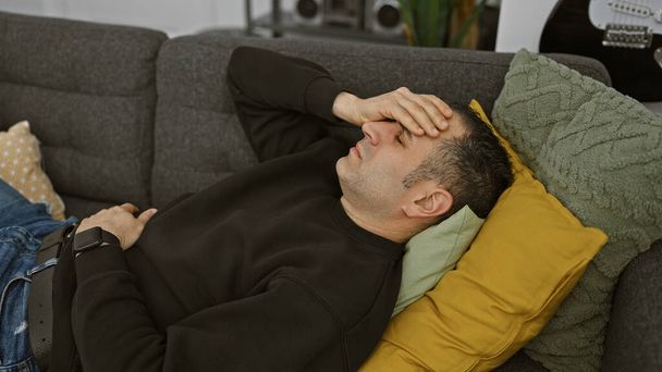 Наголошений молодий іспаномовний чоловік лежить на дивані в приміщенні, рука на лобі, виражаючи виснаження або головний біль. - Фото, зображення