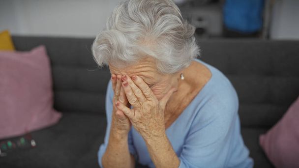 Eine besorgte ältere Frau mit grauen Haaren, die ihr Gesicht mit den Händen bedeckt, während sie drinnen auf einer Couch sitzt. - Foto, Bild