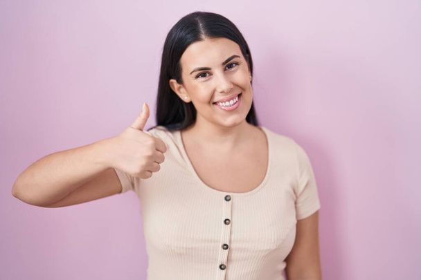 Молодая латиноамериканка, стоящая на розовом фоне, делает счастливый жест вверх рукой. одобрение выражения лица, смотрящего на камеру и показывающего успех.  - Фото, изображение