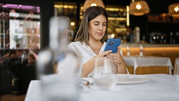 Junge schöne hispanische Frau lächelt glücklich mit Smartphone auf dem Tisch im Restaurant - Foto, Bild