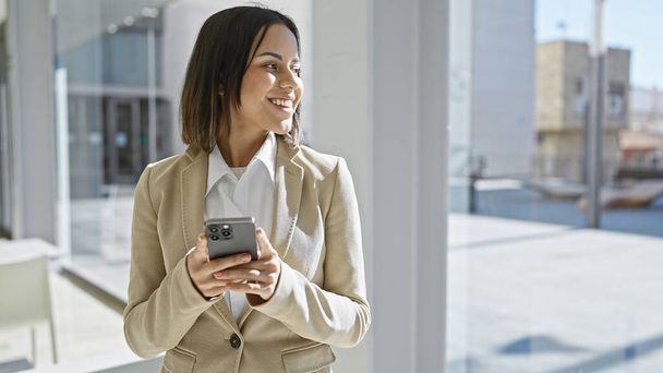 Glimlachende Spaanse vrouw in zakelijke kleding met behulp van een smartphone in een modern kantoor met een raam en stadsgezicht - Foto, afbeelding