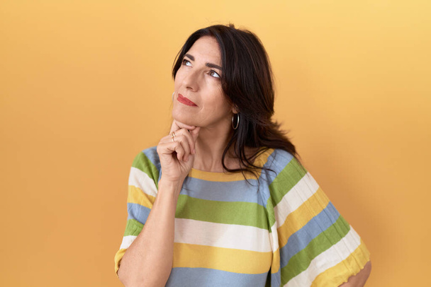 Middle age hispanic woman standing over yellow background with hand on chin thinking about question, pensive expression. smiling with thoughtful face. doubt concept.  - Fotoğraf, Görsel