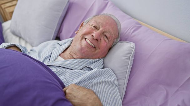 Впевнений старший чоловік, з сердечною посмішкою, насолоджується затишним ранком, лежачи в ліжку в інтимній атмосфері своєї комфортної спальні. - Фото, зображення