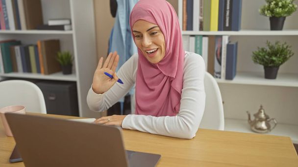 Mulher sorridente em hijab usando laptop em casa, acenando durante uma chamada de vídeo em um interior moderno. - Foto, Imagem