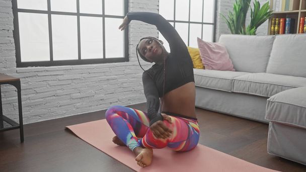 Afroamerykanka rozciągająca się na macie do jogi w nowoczesnym salonie z sofą w tle. - Zdjęcie, obraz