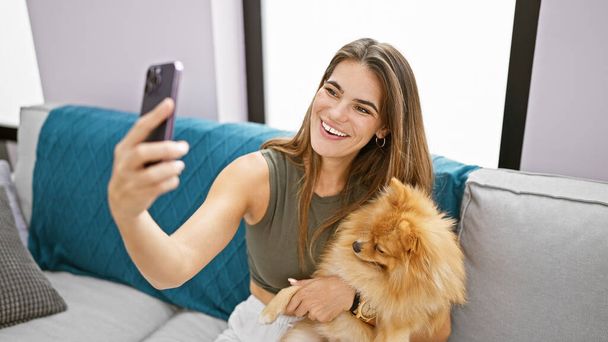 Vidám, fiatal spanyol nő kényelmesen ül otthon a nappali kanapéján egy mosollyal. élvezi a beltéri időt, hogy egy boldog szelfi kép vele kisállat kutya segítségével smartphone technológia. - Fotó, kép