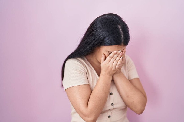 Jonge Spaanse vrouw die over een roze achtergrond staat met droevige uitdrukking en gezicht bedekt met handen terwijl ze huilt. depressie concept.  - Foto, afbeelding