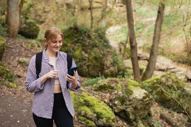 Girl tourist walking in forest and using smartphone. Young female with backpack hiking on the road in nature. Beautiful 30s woman walking on nature in sport clothes - Photo, Image