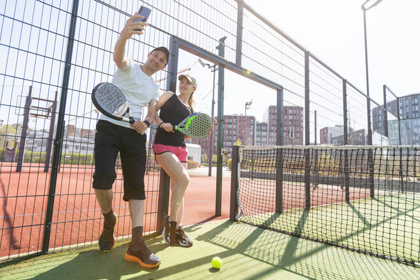 Retrato de mulher jovem positiva e homem adulto em pé na quadra de tênis padel, segurando raquete e bola, sorrindo. Foto de alta qualidade - Foto, Imagem