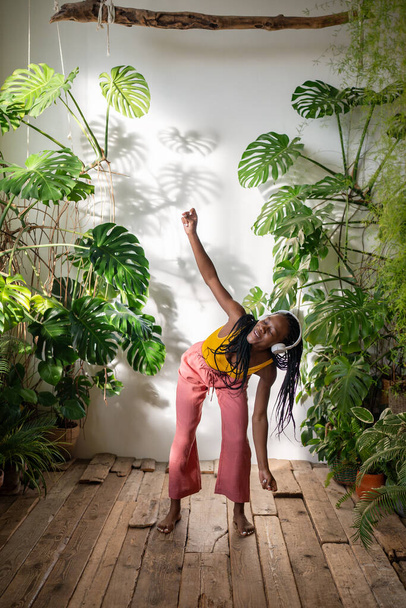 Безтурботна чорна жінка танцює босоніж в навушниках, слухаючи музику на дерев'яній підлозі із задоволенням, насолоджуючись моментом. Розслаблена афроамериканська молода жінка, що охолоджує пісні в кімнаті з домашніми рослинами - Фото, зображення