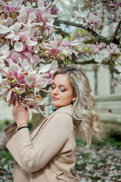Belle jeune blonde avec bouquet de fleurs. Portrait de jeune femme près de magnolia en fleurs dans la ville européenne. Printemps. - Photo, image
