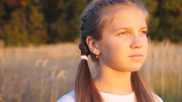 Portrait of happy little girl stands at grass meadow looking at beautiful evening sunset. Cute female face illuminating by bright rays of sun at summer day. Carefree and freedom concept. Close up. - Záběry, video