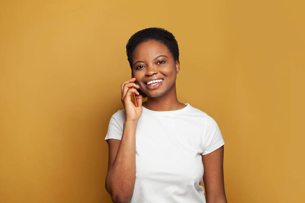 Alegre mujer afroamericana hablando por teléfono en el fondo de la bandera de color amarillo brillante - Foto, imagen