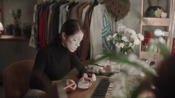Νεαρή ηθοποιός με mime μακιγιάζ που απλώνει πούδρα προσώπου με πινέλο ενώ κάθεται στο γραφείο ματαιοδοξίας στο καμαρίνι και ετοιμάζεται για παράσταση - Πλάνα, βίντεο