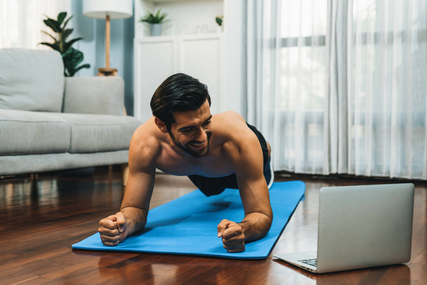 Athletic and sporty man doing plank on fitness mat during online body workout exercise session for fit physique and healthy sport lifestyle at home. Gaiety home exercise workout training. - Photo, Image
