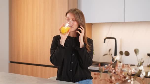 Boldog mosolygós fiatal nő a konyhában beszél okostelefonon és iszik pohár narancslevet. - Felvétel, videó