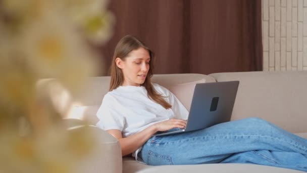 Giovane donna seduta sul divano in soggiorno e utilizzando il suo computer portatile. - Filmati, video