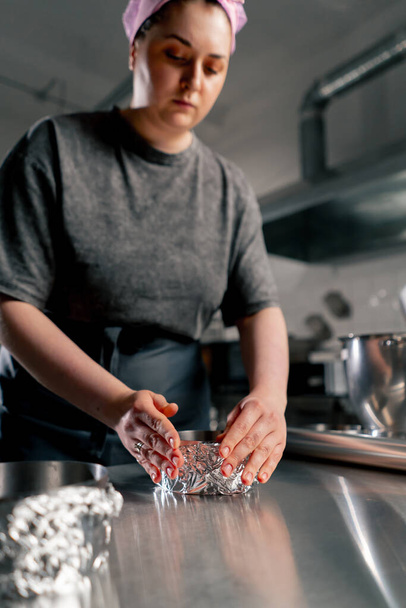 χαμηλή βολή θηλυκό αρτοποιός σε μια επαγγελματική κουζίνα τυλίγει τα τηγάνια με αλουμινόχαρτο πριν από τη ρίψη της ζύμης πάνω στο κέικ - Φωτογραφία, εικόνα