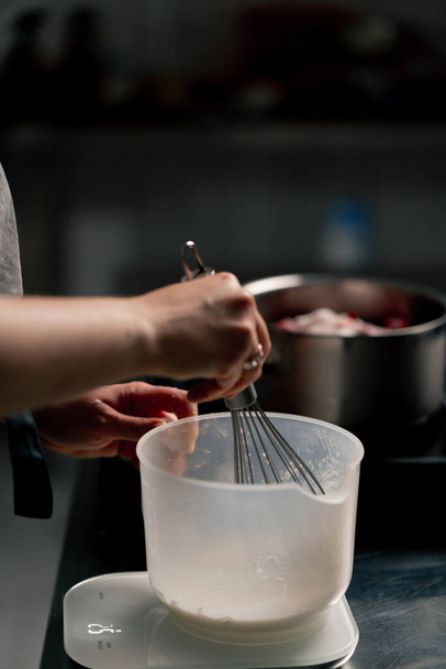 primer plano del panadero en una cocina profesional batiendo masa en un recipiente transparente con un batidor de mano - Foto, imagen