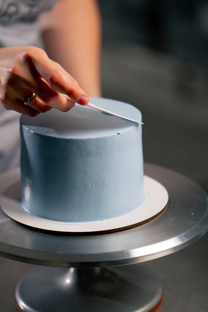 close up padeiro feminino em uma cozinha profissional distribui creme azul em um bolo de esponja com uma espátula - Foto, Imagem