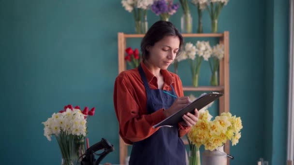 people, business and floristry concept. Portrait of happy smiling florist woman making notes to clipboard at her flower shop, looking at camera with smile. Copy space - Footage, Video