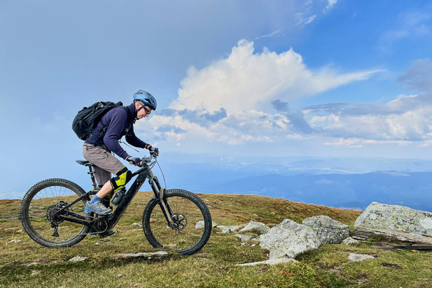 Ποδηλάτης με ηλεκτρικό ποδήλατο βουνού σε εξωτερικούς χώρους. Αρσενικό τουριστικό ποδήλατο κατά μήκος χορτώδες μονοπάτι στα βουνά, φορώντας κράνος και σακίδιο πλάτης. Έννοια του αθλητισμού, της ενεργούς αναψυχής και της φύσης. - Φωτογραφία, εικόνα