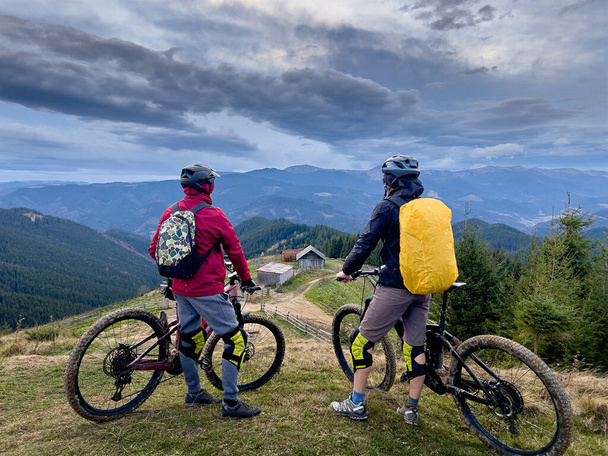 Δύο ποδηλάτες οδηγούν ηλεκτρικά ποδήλατα έξω. Επιστροφή άποψη των ανδρών τουριστών στηρίζεται στην κορυφή του λόφου, απολαμβάνοντας όμορφο ορεινό τοπίο, φορώντας κράνος και σακίδιο πλάτης. Έννοια του ενεργού ελεύθερου χρόνου. - Φωτογραφία, εικόνα