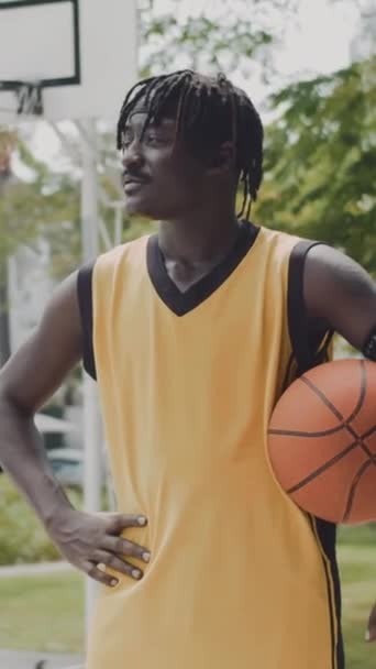裁判所の屋外でカメラで微笑む肯定的なアフリカ系アメリカ人のバスケットボール選手の垂直な肖像画 - 映像、動画