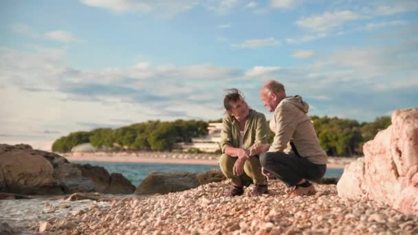 vacaciones para los pensionistas, ancianos encantador hombre y mujer se divierten lanzando guijarros en el mar mientras se sienta en la orilla durante el viaje turístico para las personas mayores - Imágenes, Vídeo