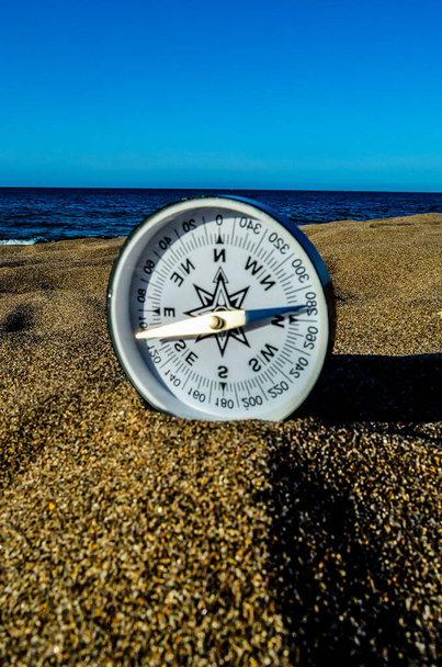 Uma bússola está sentada na areia, apontando para o norte. A imagem tem um clima calmo e tranquilo, como a bússola é colocada em uma praia com o oceano no fundo - Foto, Imagem