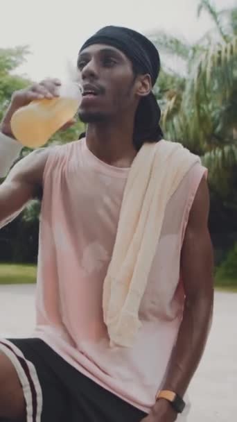 Retrato vertical de un atleta negro bebiendo limonada fresca y sonriendo a la cámara después del entrenamiento al aire libre - Imágenes, Vídeo