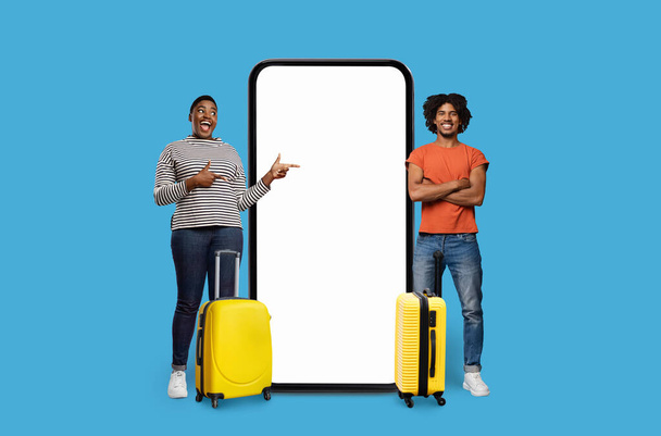 Ένας κατάπληκτος μαύρος και ένας σίγουρος άνθρωπος στέκονται από ένα υπερμεγέθης τηλέφωνο με μια κενή οθόνη για διαφήμιση εφαρμογή, απομονώνονται σε ένα ζωντανό μπλε φόντο - Φωτογραφία, εικόνα