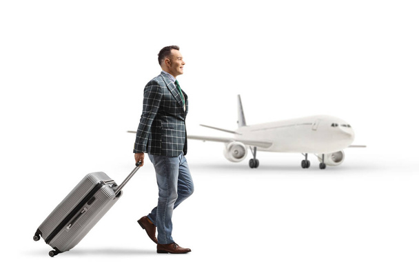 Full length profile shot of a man in suit and jeans walking and pulling a suitcase at the airport isolated on white background - Valokuva, kuva