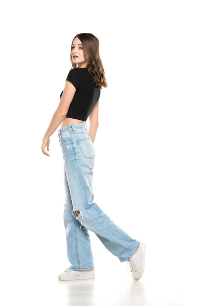 Giovane modella donna con jeans strappati e camicia nera che cammina su uno sfondo bianco da studio. Vista laterale, profilet - Foto, immagini