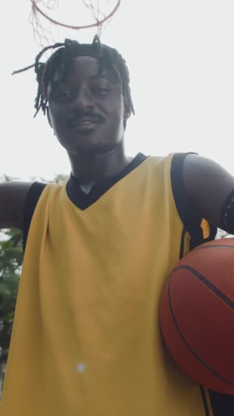 Retrato vertical de bajo ángulo del alegre deportista negro mirando a la cámara sosteniendo la pelota cerca del aro de streetball - Metraje, vídeo