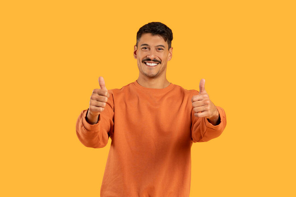 Een duizendjarige man met een snor en een grappige uitdrukking geeft twee duimen omhoog in een stralende oranje trui, geïsoleerd tegen een oranje achtergrond - Foto, afbeelding