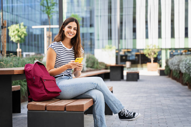 Estudiante hispana usando aplicación de teléfono celular amarillo en el campus de la Universidad, mirando a la cámara con sonrisa positiva - Foto, Imagen