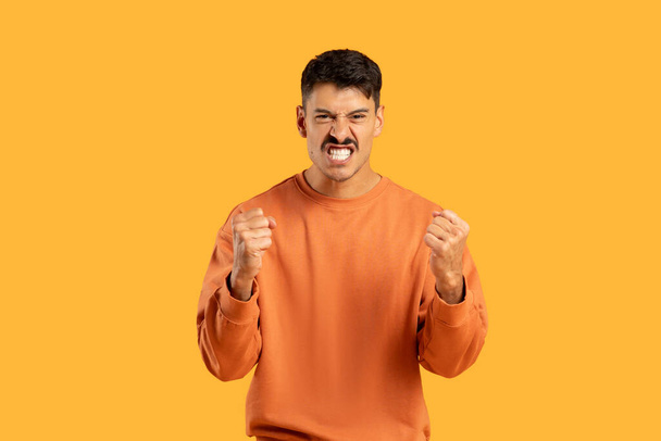 Un giovane uomo che mostra un forte gesto di fastidio o rabbia, stringendo i pugni saldamente su uno sfondo arancione chiaro - Foto, immagini
