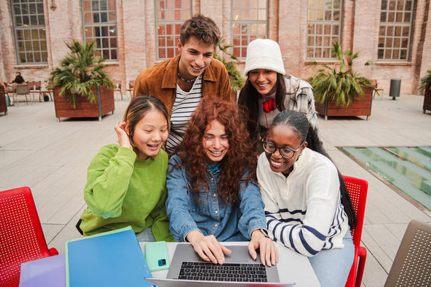 Bir grup neşeli genç, üniversite kampüsünde toplanmış laptoplarını kullanarak bilgisayardan bilgi toplamaya odaklanmış bir ödev üzerinde çalışıyorlar. - Fotoğraf, Görsel