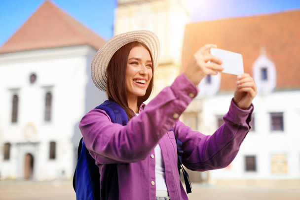 Femme souriante avec chapeau de soleil prenant un selfie avec son téléphone avec des bâtiments historiques derrière, concept de tourisme - Photo, image