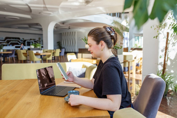 Mujer joven enfocada utiliza simultáneamente la tableta y el ordenador portátil en un espacio de coworking brillante y lleno de plantas. - Foto, imagen