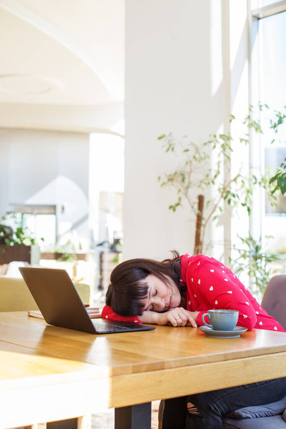 Wyczerpana młoda bizneswoman śpiąca przy biurku z laptopem i filiżanką kawy w słonecznym, ozdobionym roślinami biurze. - Zdjęcie, obraz