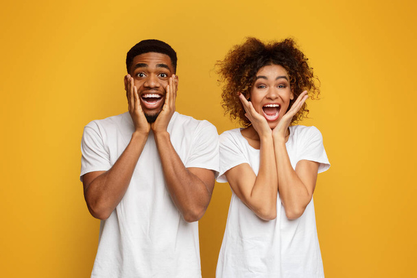 African-american woman and man look joyfully at camera, keep hands on cheeks, express positive feeling and amazement, orange background - Photo, Image
