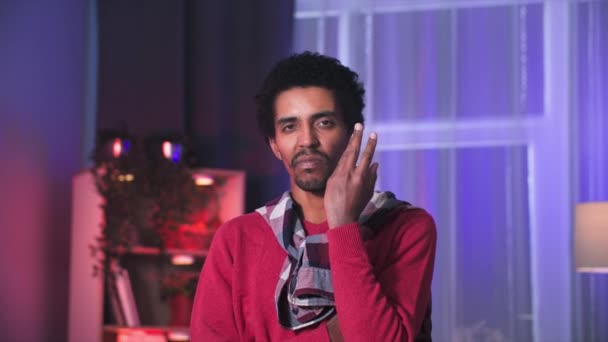 jonge zwarte man aanraken van zijn gezicht met de handen en bedachtzaam te kijken naar de camera terwijl staan in de kamer - Video