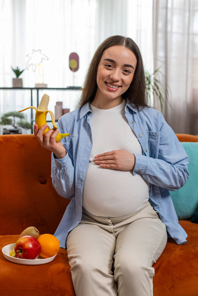 Mujer embarazada feliz con gran apetito pela y come plátano fruta se sienta en el sofá en la sala de estar con interior moderno. Cuidado de la maternidad cuidado de los niños. Chica sonriente elige la comida sana correcta. Vertical - Foto, Imagen