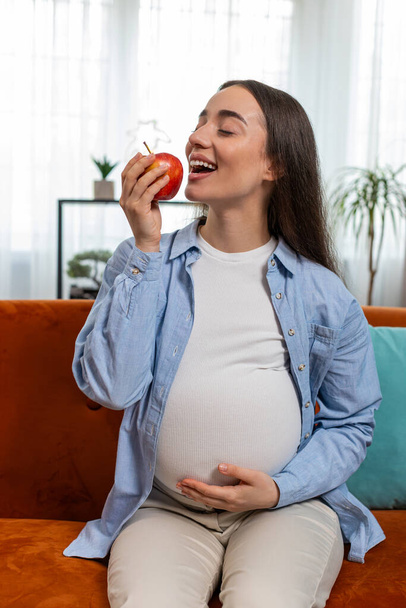 Donna incinta felice mangiare frutta fresca mela rossa seduta sul divano in salotto interno. Maternità, cura di sé, assistenza all'infanzia. Donna sorridente guardando la fotocamera sceglie giusto cibo vitaminico sano. Verticale - Foto, immagini