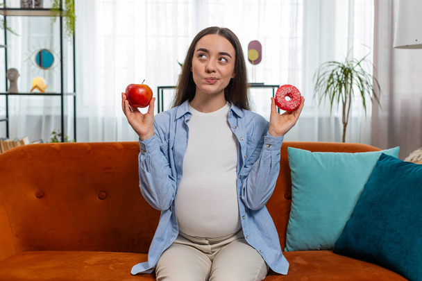 Choosing right nutrition. Confused young pregnant woman comparing sweet donut and ripe apple and shrugging shoulders in uncertainty. Future mother girl hesitates choosing between dessert and fruit. - Photo, Image