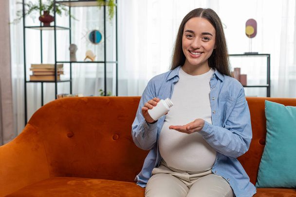 Pregnant Caucasian woman taking drug and drinking water to nourish unborn child sitting on sofa couch in living room at home. Health care medical and pregnancy concept. Lady happily looks at camera. - Photo, Image