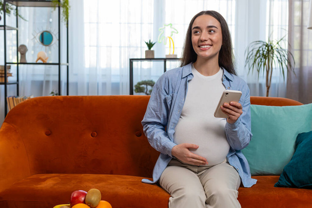 Sourire femme enceinte en utilisant la messagerie texte smartphone médias sociaux bavardage assis sur le canapé dans le salon à la maison. Heureuse fille caucasienne future mère caresser le ventre faire des achats en ligne pour bébé - Photo, image